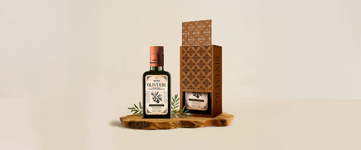 Award-winning premium olive oil packaging design for Olivixir Nikolos Collection by ABC Design, featuring an elegant dark glass bottle with heritage-style typography alongside a luxury laser-cut wooden gift box on a rustic wood base.