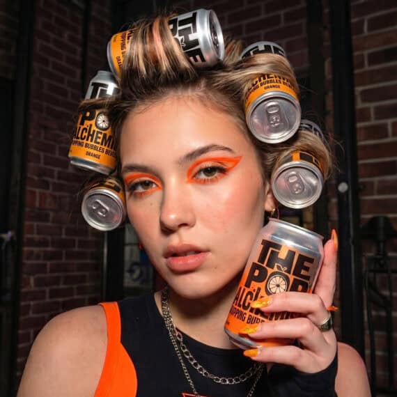 Close-up of a woman with orange makeup using Pop Alchemist cans in her hair, beverage design by ABC Design Communication.